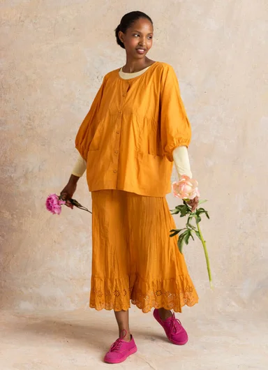 A woman holding flowers, dressed in a matching orange blouse and skirt in woven fabric from Gudrun Sjödén.