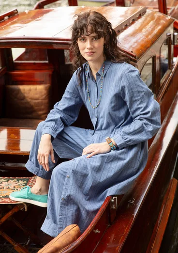 Robe tissée bleue à rayures avec manches longues et boutons. Combinée avec un pantalon bleu et des chaussures vert-bleu.
