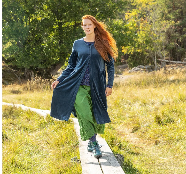 A woman on a boardwalk in an autumnal setting wearing blue velour, a patterned blouse and green trousers from Gudrun Sjödén.