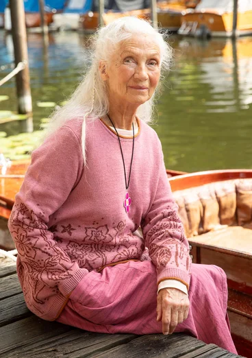 Eine Dame sitzt am Bootssteg mit roséfarbenen Pullover. Der untere Teil des Oberteils hat Blumen aufgestickt. Untenrum trägt die Dame einen gestreiften Rock in altrosé.  