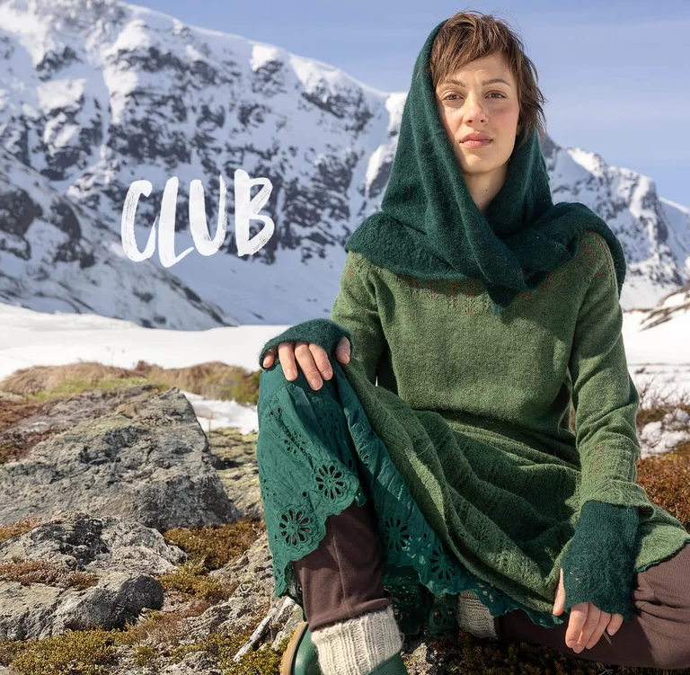 A woman sitting in front of a snowy mountain wearing layers of green clothes from Gudrun Sjödén’s 2025 Christmas collection.