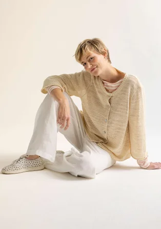 Model trägt eine beige Strickjacke mit Knöpfen. Unter der Strickjacke trägt die Frau eine weiß-beiges Streifenshirt. Außerdem hat die Dame eine weiße Hose an. 