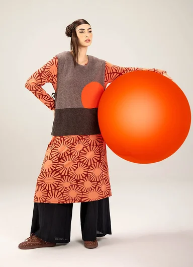 A woman who seems to be holding a large, orange ball, wearing layers of clothes from Gudrun Sjödén.