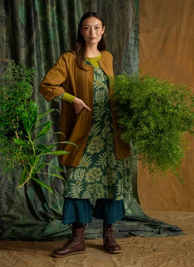 A woman holding a large green plant wearing layers of clothes from Gudrun Sjödén.