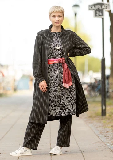 Robe tissée noire à rayures, portée ouverte et avec des manches longues. En dessous, le modèle porte une robe noire et blanche avec une ceinture rouge. Combinée avec un pantalon noir à rayures et des chaussures blanches à pois. 
